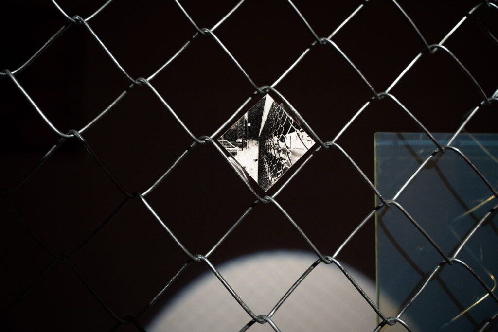 documentation of installation showing people looking at a hologram and pinhole photos in a chain link fence at the exhibition Middles. photos by Jian Peters